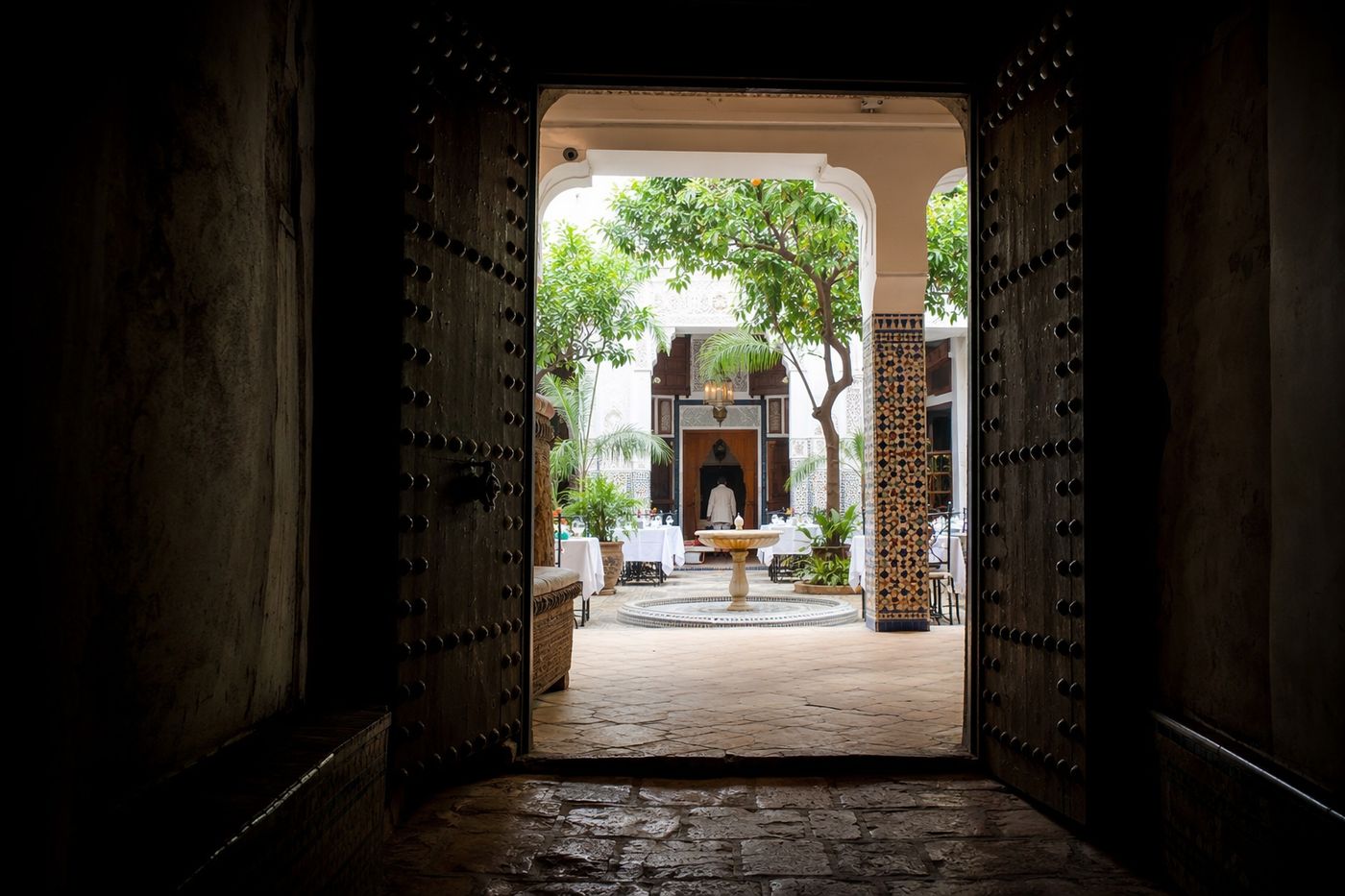 Patio lumineux d'un riad-restaurant aperçu depuis un passage sombre, à travers une lourde porte cloutée