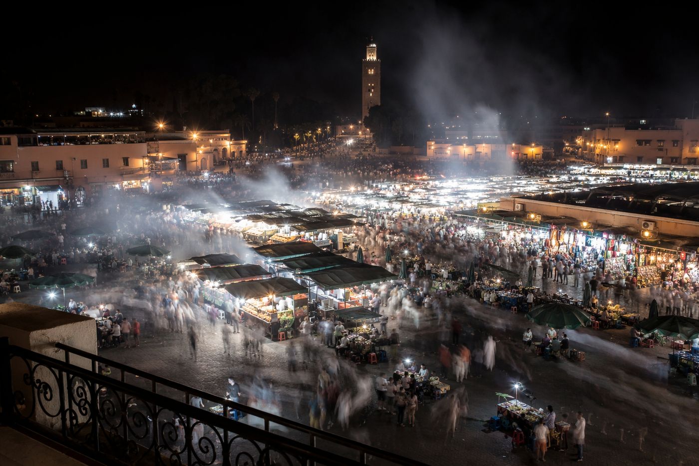 Grande place vue de nuit depuis une terrasse, stands fumants sous des ampoules blanches et foule en mouvement