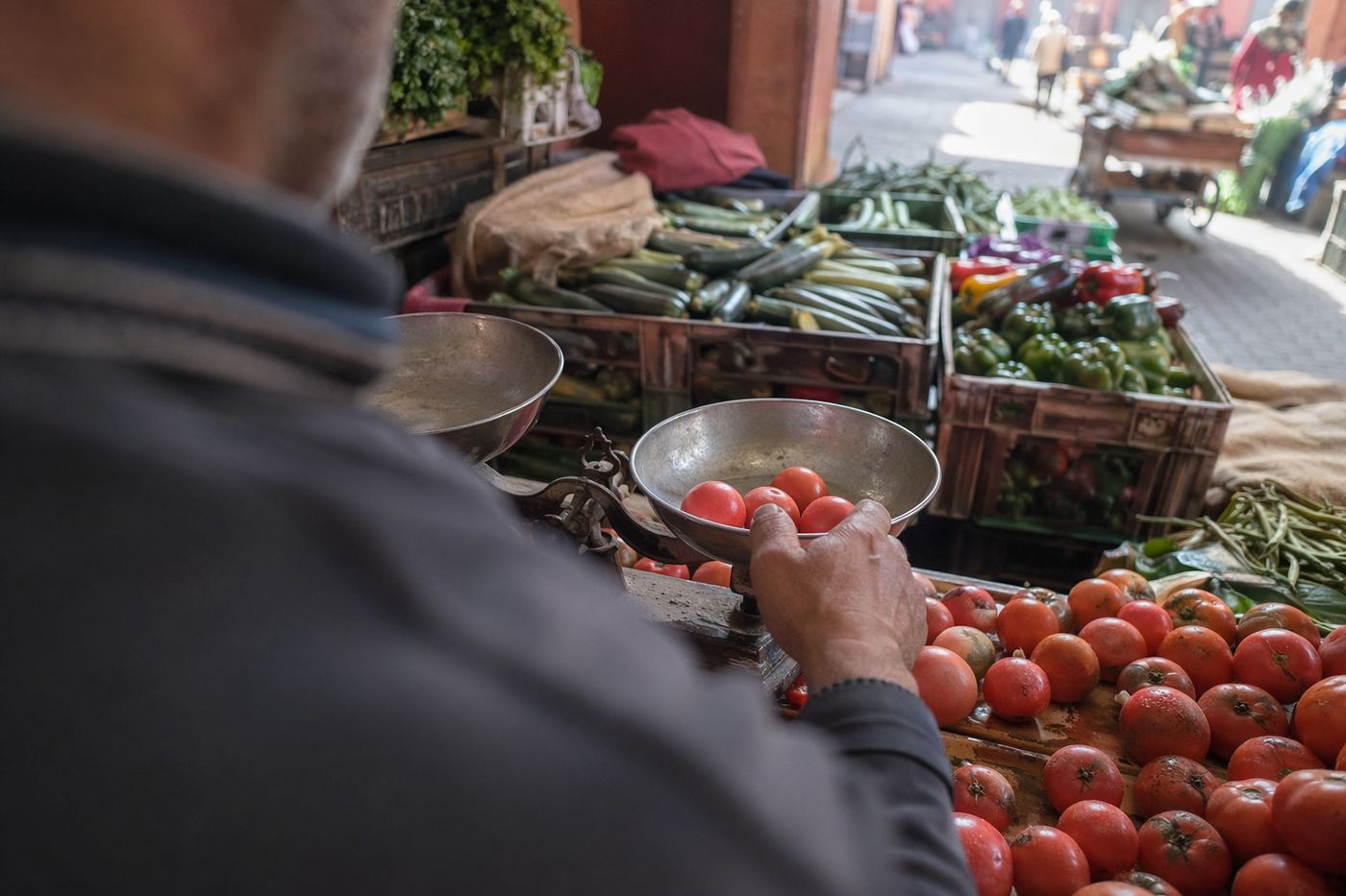 Mains d'un marchand pesant des tomates sur une balance à plateaux, caisses de légumes derrière.
