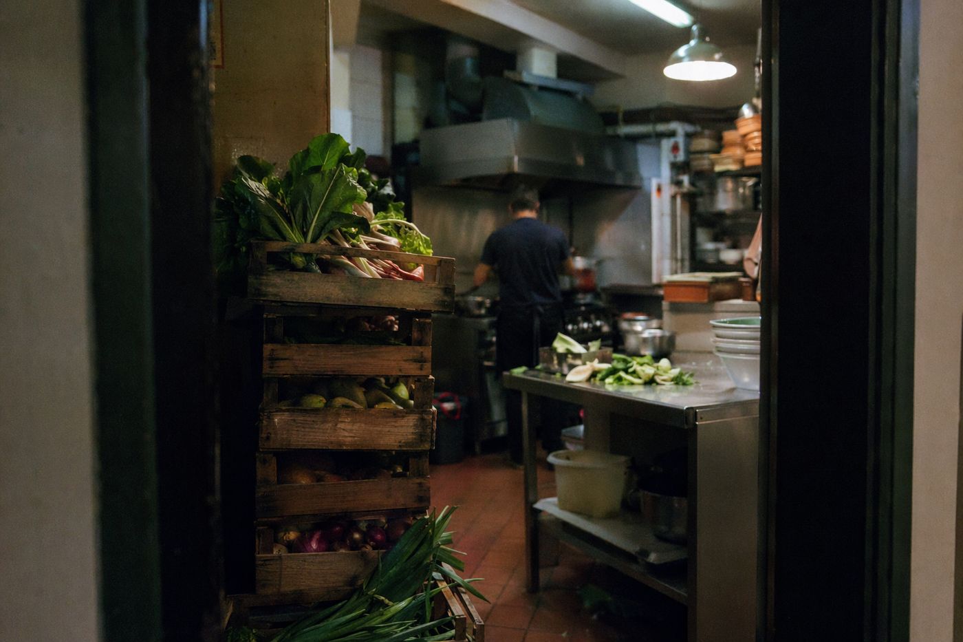 Cuisine de restaurant vue depuis la porte, caisses de légumes du marché empilées et cuisinier de dos.