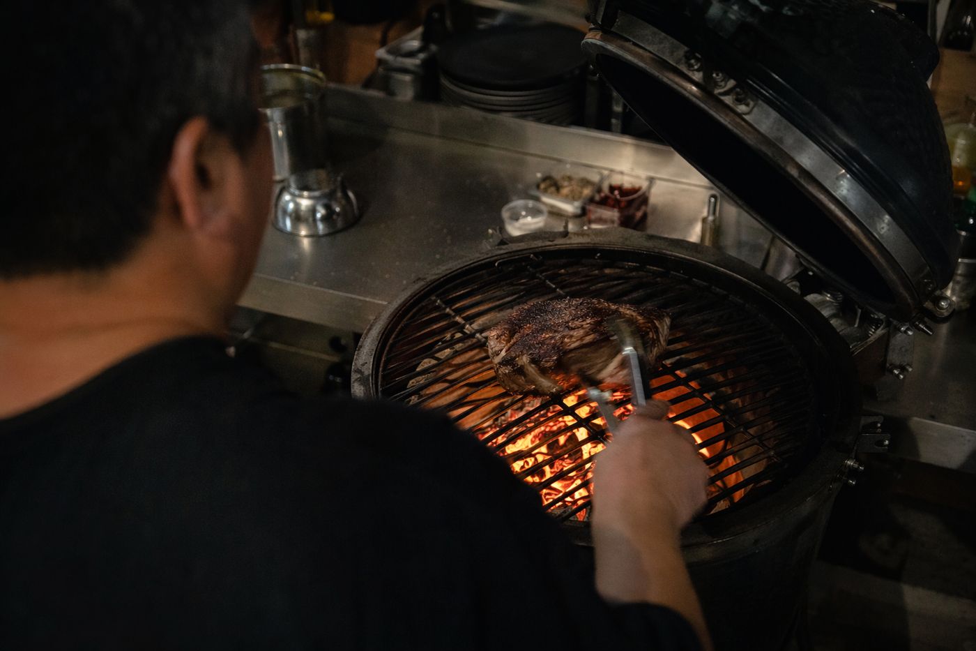Mains d'un cuisinier retournant une grosse pièce de bœuf sur un gril au charbon, braises rougeoyantes en dessous
