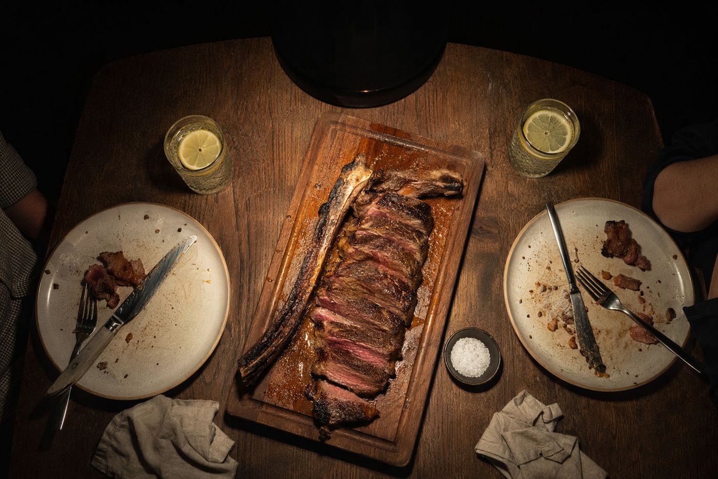 Côte de bœuf tranchée sur planche entre deux assiettes et deux verres d'eau pétillante, sous une suspension basse