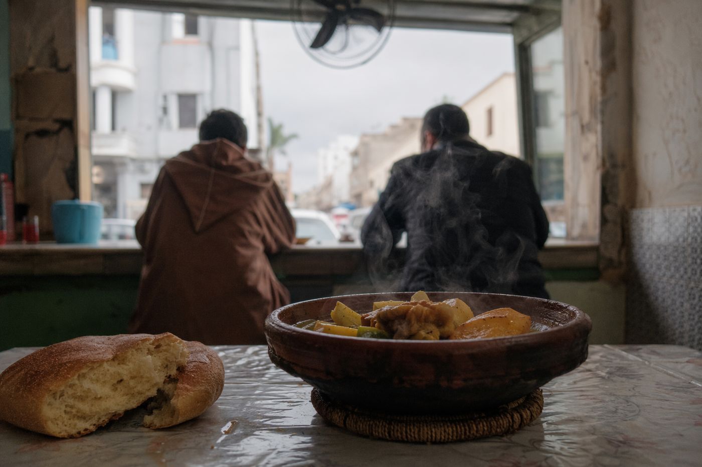 Tajine fumant sur la table d'une petite cantine de quartier, clients attablés au fond, de dos