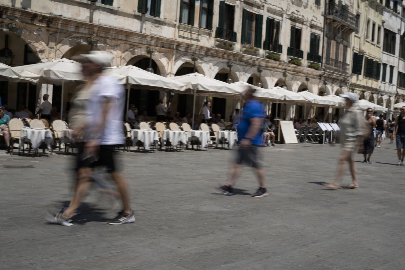Rangée de terrasses à touristes alignées sous parasols au bord d'une grande place, plein midi
