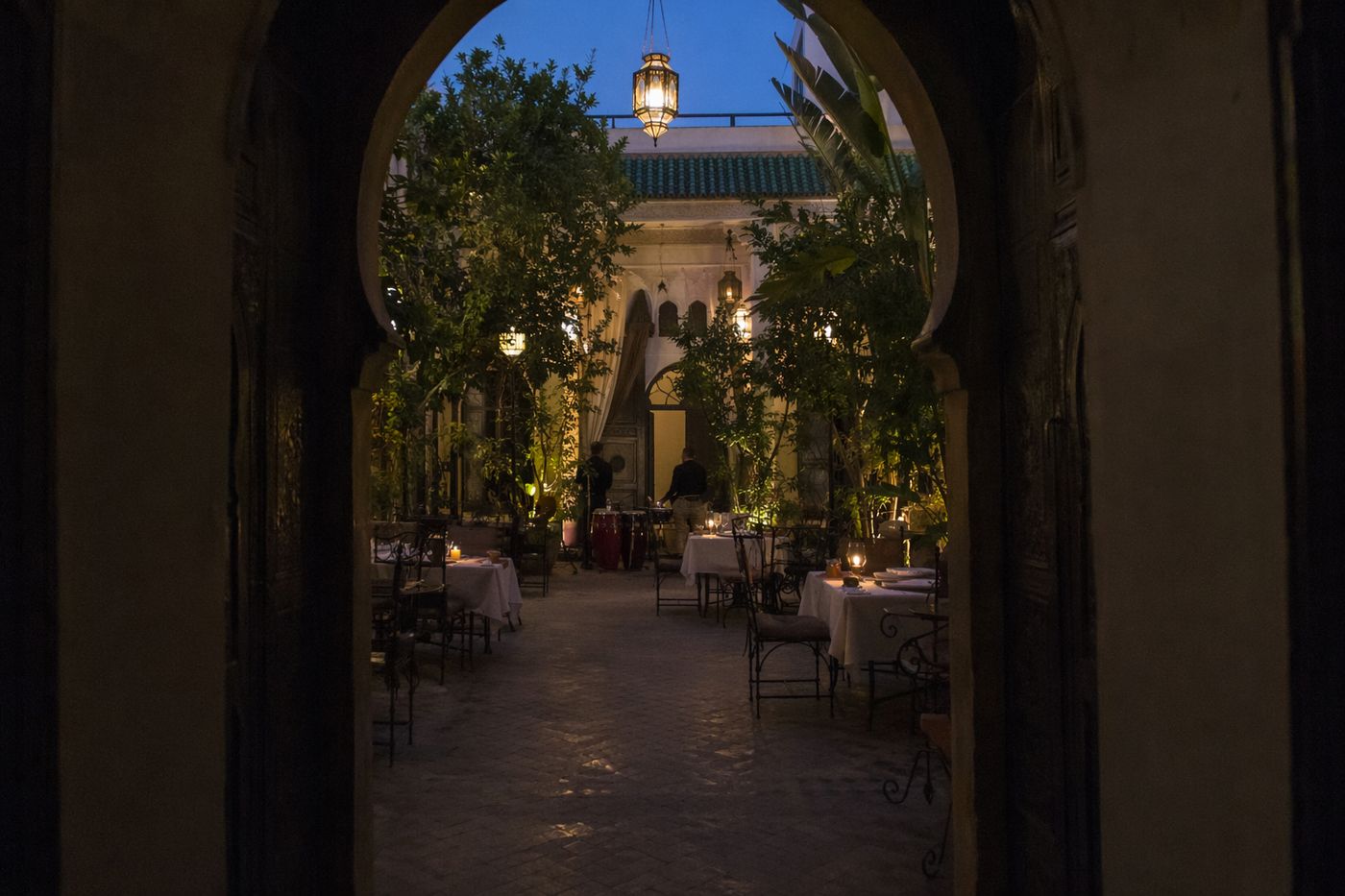 Cour de riad à l'heure bleue vue d'une arche, lanternes allumées et musiciens installant leurs tambours.