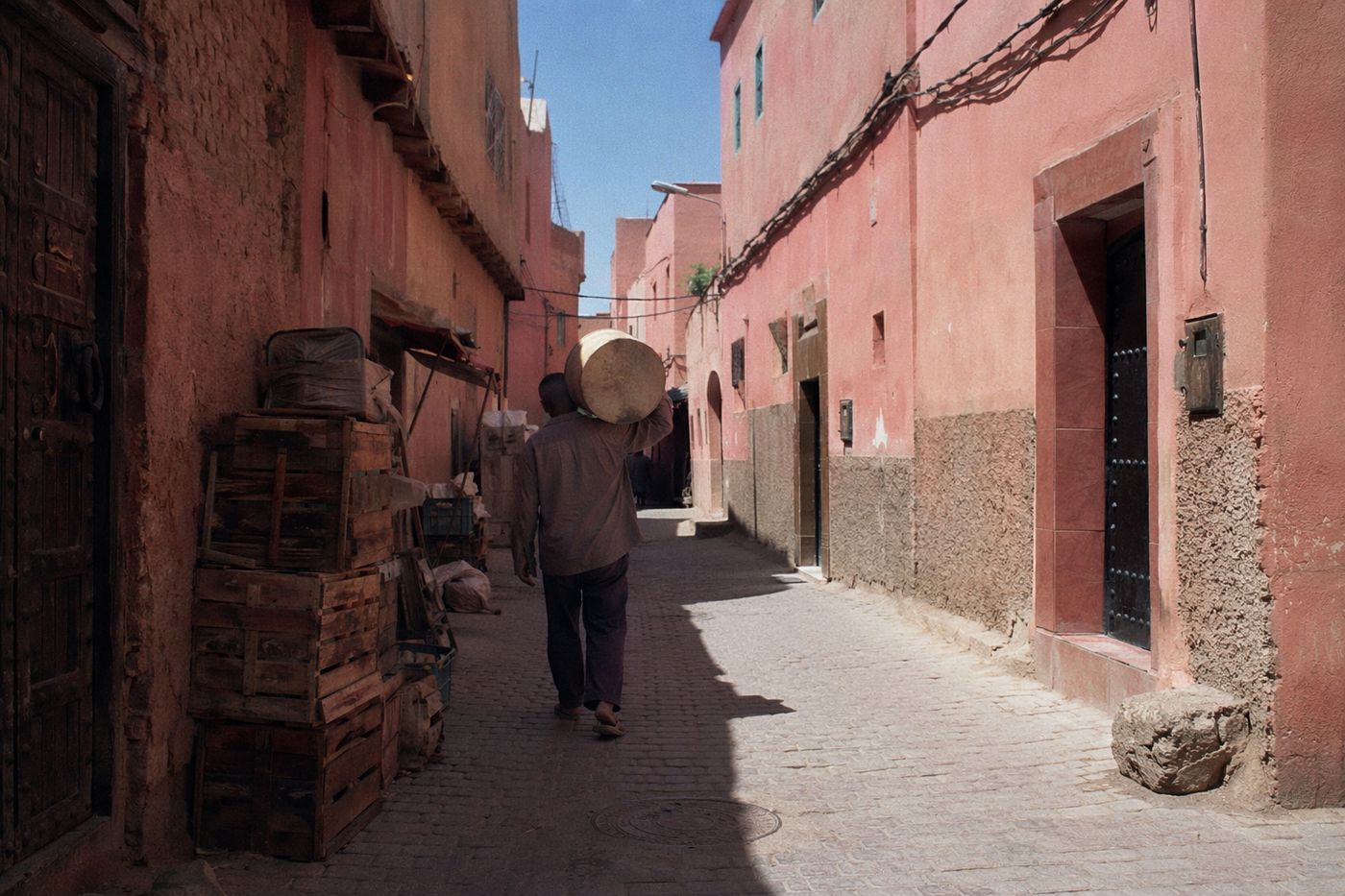 Ruelle de la kasbah sous le soleil de midi, silhouette portant un tambour s'éloignant.