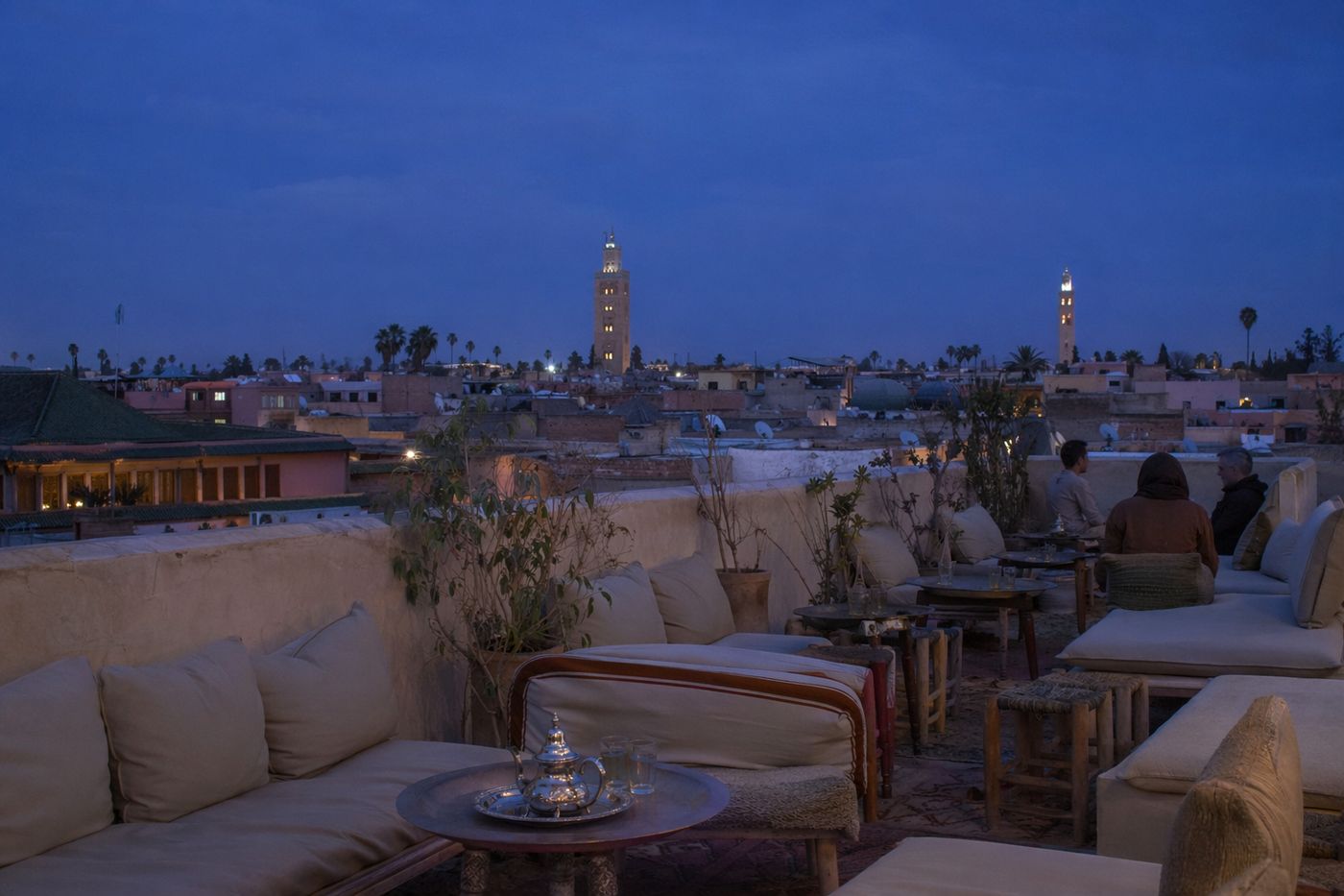 Terrasse sur les toits au crépuscule, théières et verres sur les tables basses, minarets dans le ciel bleu.