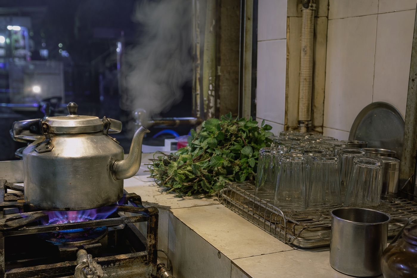 Comptoir de café le soir : théière cabossée sur brûleur à gaz, tas de menthe fraîche et verres retournés.