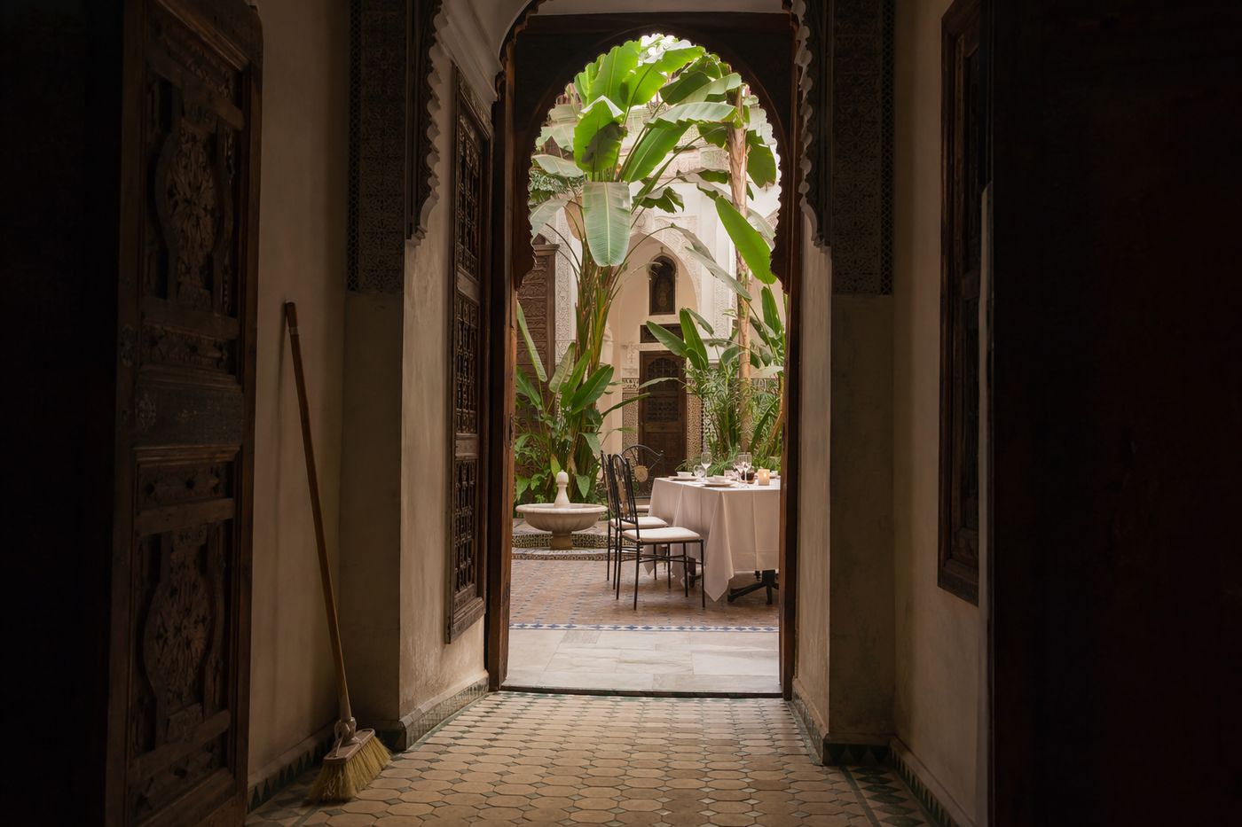 Cour plantée d'un riad vue depuis un couloir sombre, fontaine et table dressée dans la lumière.