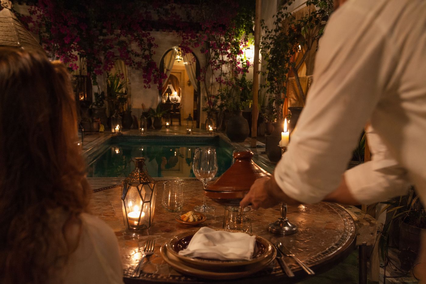 Mains d'un serveur posant un tajine sur une table aux bougies dans un patio de riad, le soir.