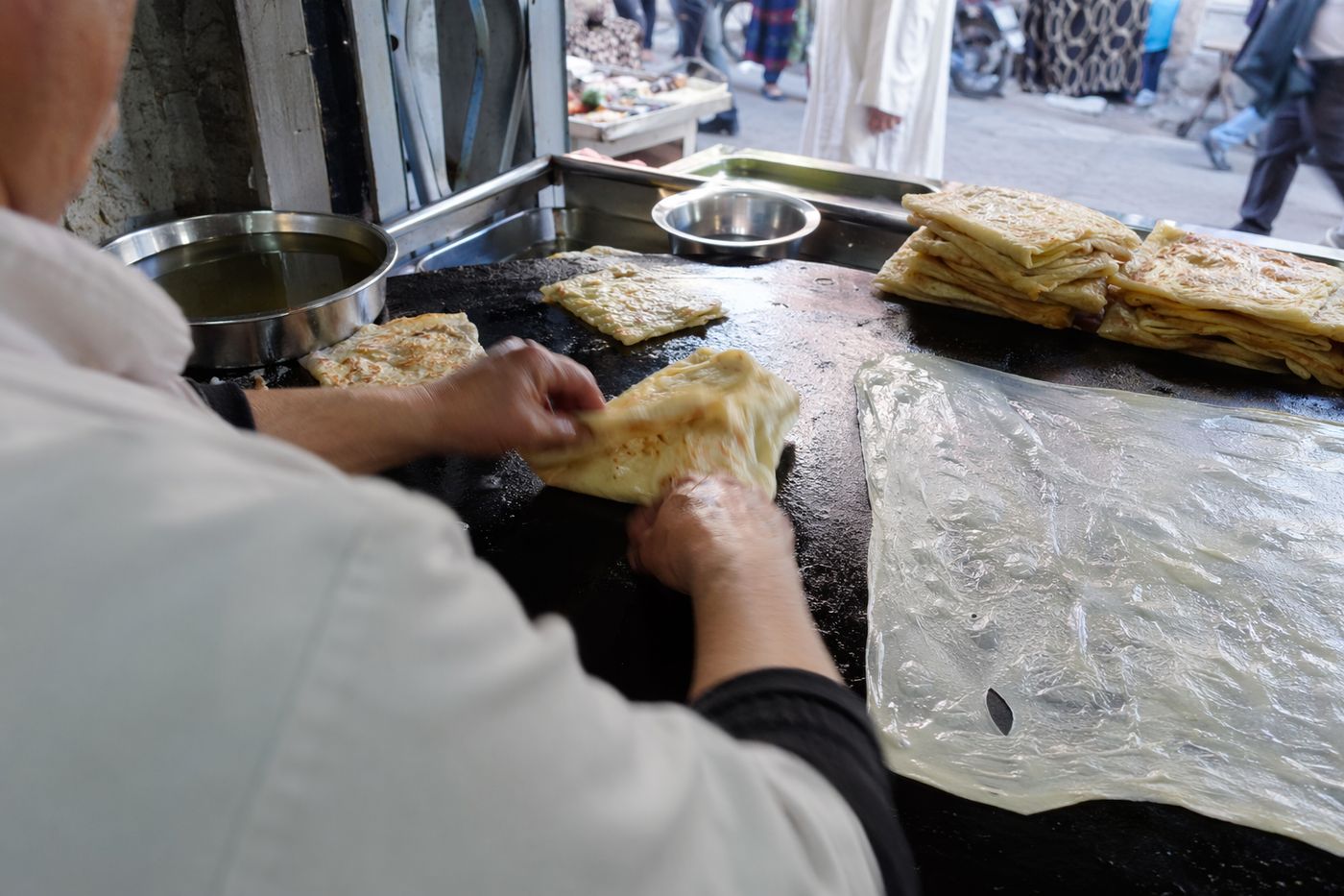 Mains d'un cuisinier pliant un msemen sur une plaque, pâte étirée et pile de crêpes dorées à côté.