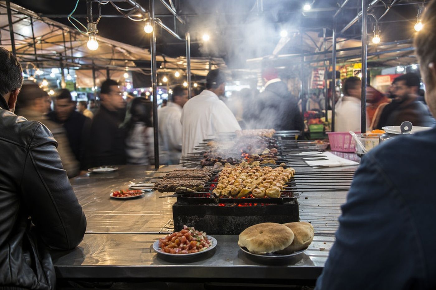 Comptoir d'un étal de grillades la nuit sur Jemaa el-Fna, brochettes au charbon, fumée et ampoules nues.
