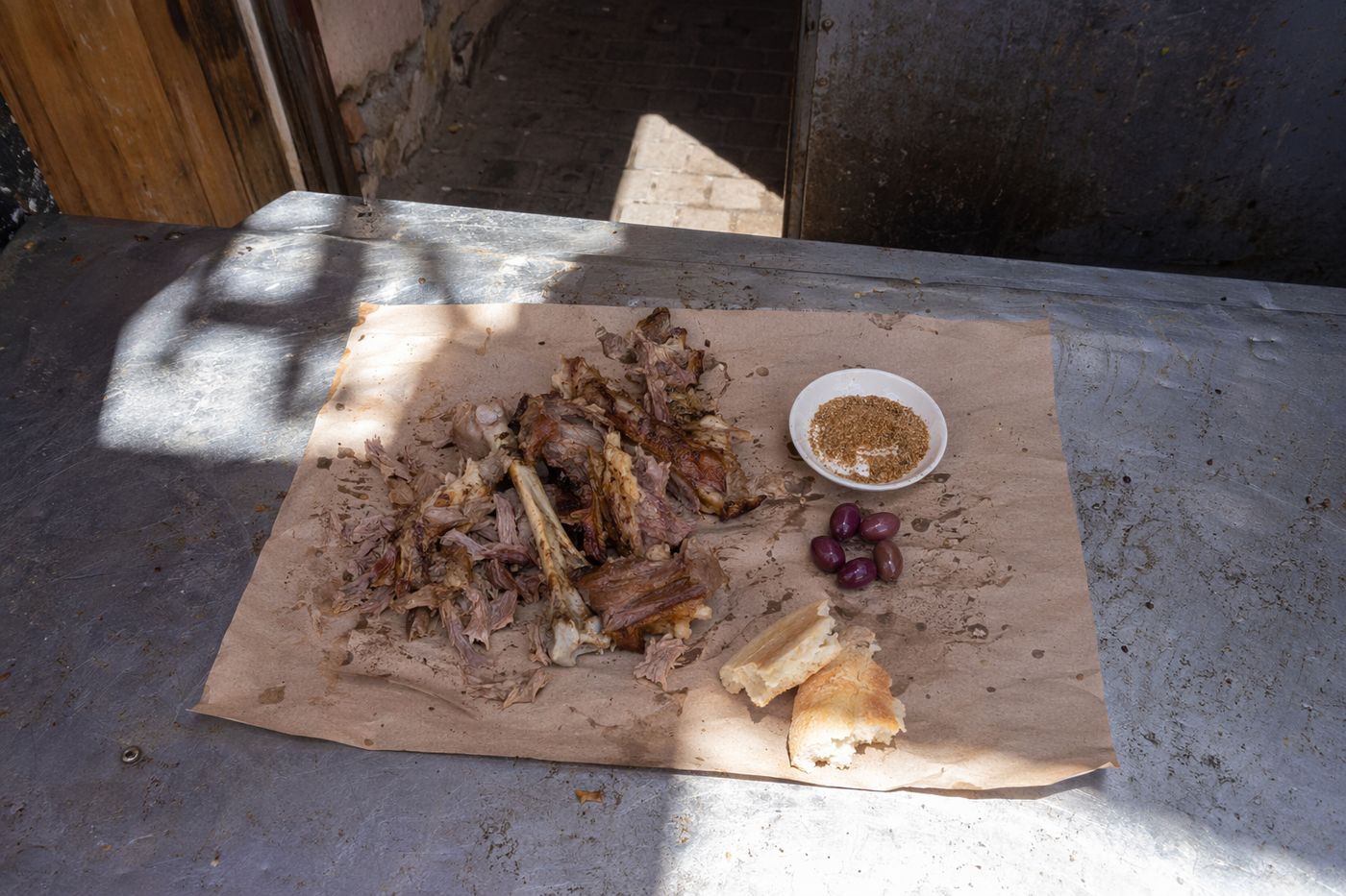 Agneau mechoui effiloché sur papier avec bol de sel-cumin, olives violettes et pain, vu du dessus à midi.