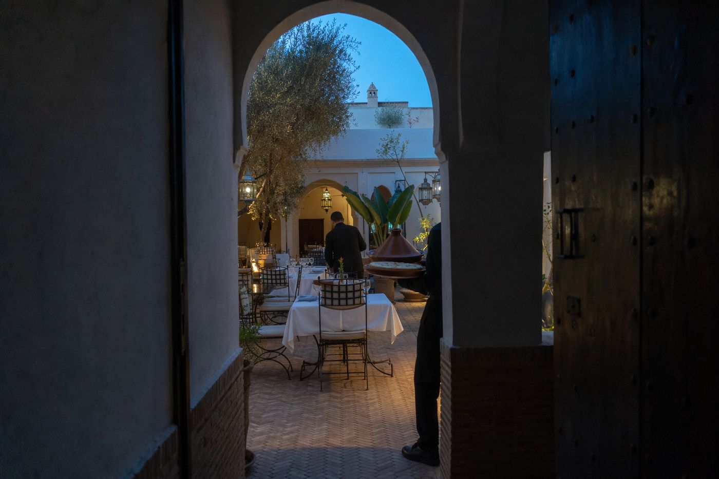 Patio au crépuscule vu à travers une arche, tables en cours de dressage et pastilla portée vers le fond.