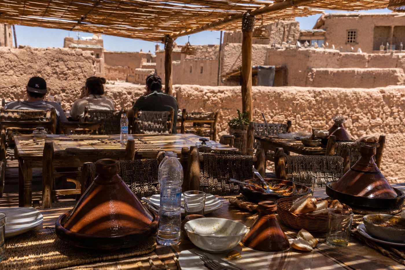 Terrasse de déjeuner rayée d'ombres dures, couvercles coniques de tajines et paniers de pain sur les tables.