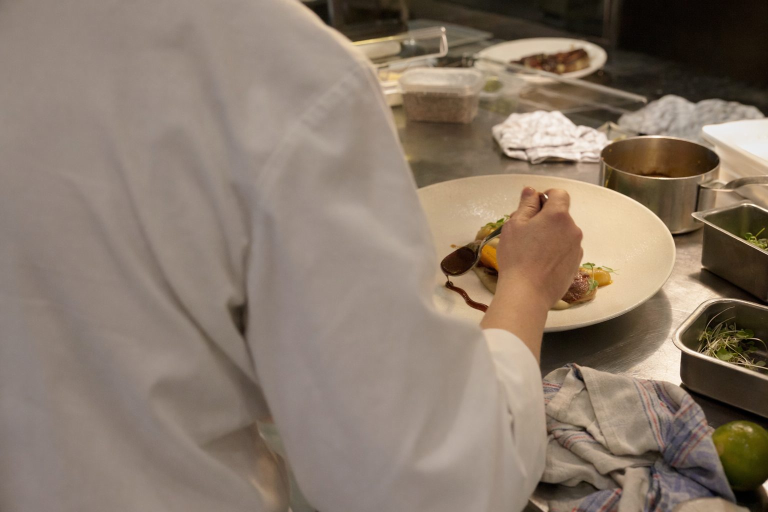 Mains d'un chef dressant une assiette au passe d'une cuisine, le soir, dos tourné à l'objectif.