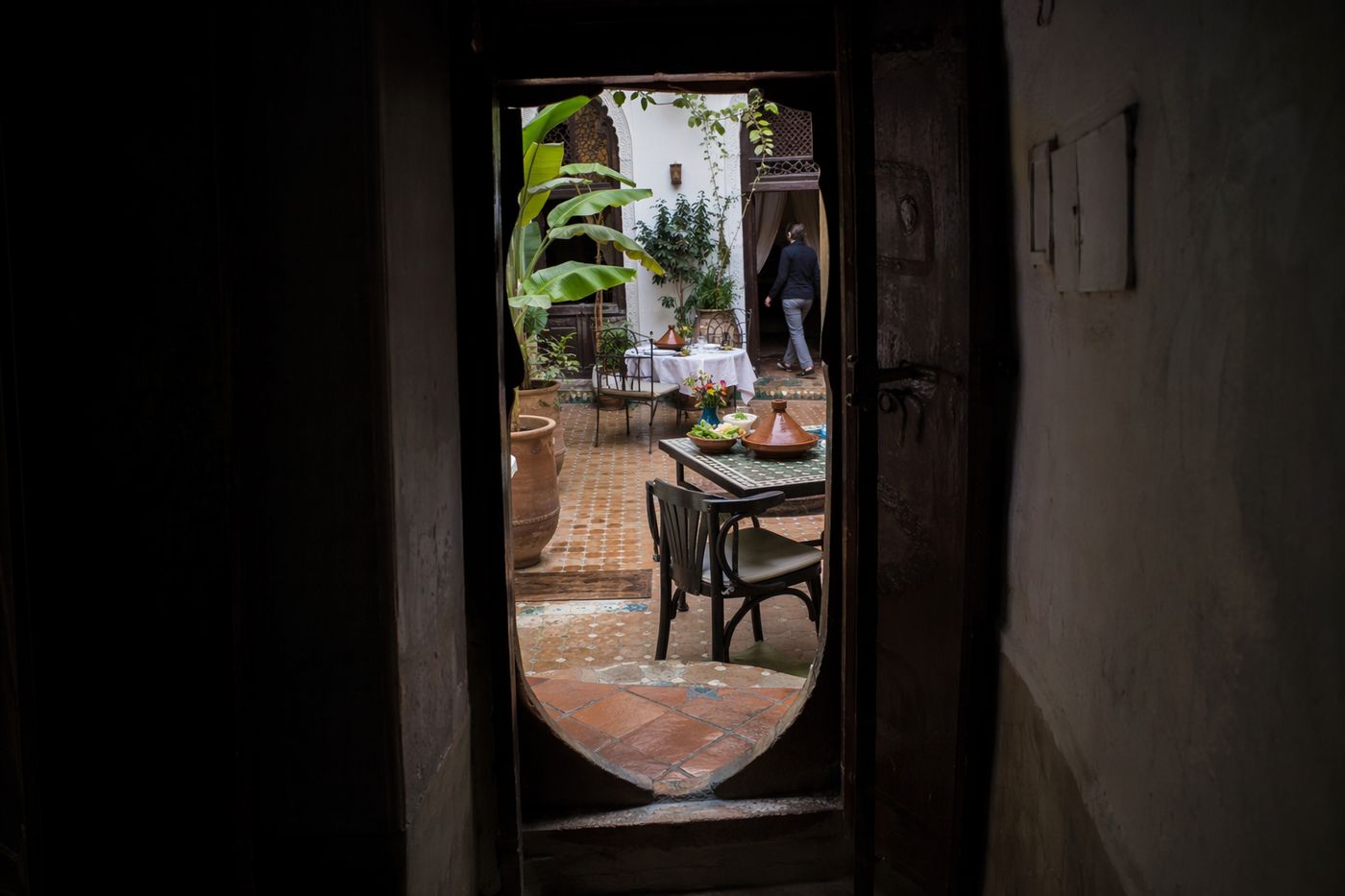 Patio de riad vu depuis un couloir sombre, tables dressées avec des tajines de légumes dans la cour lumineuse.
