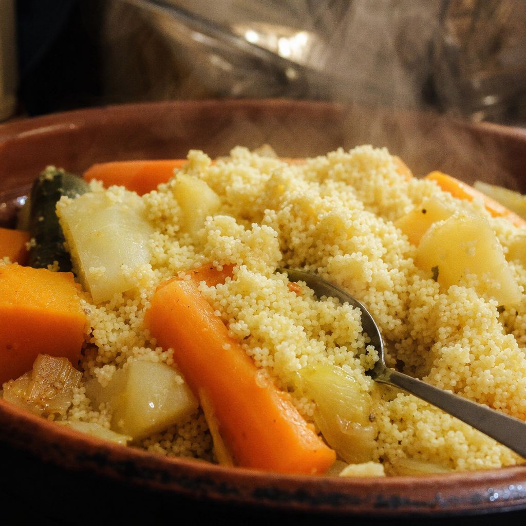 Gros plan sur des grains de couscous vapeur aux légumes dans un plat en terre, vapeur encore visible.