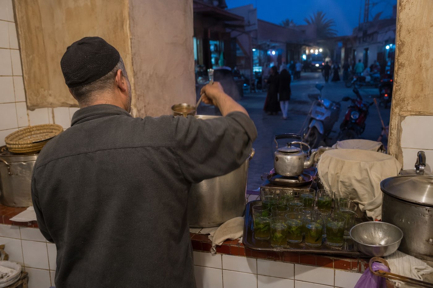 Mains d'un cuisinier servant la soupe au comptoir d'une gargote de quartier, verres de thé prêts, rue au crépuscule.